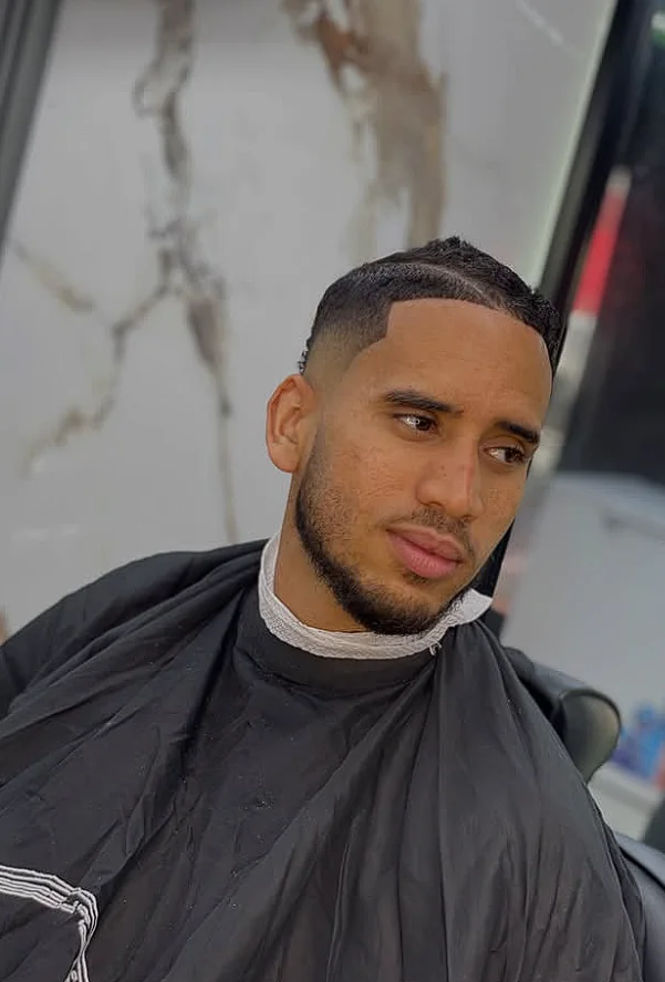 Cliente con fade bajo, raya marcada al lado y barba corta perfilada, frente a una pared de mármol